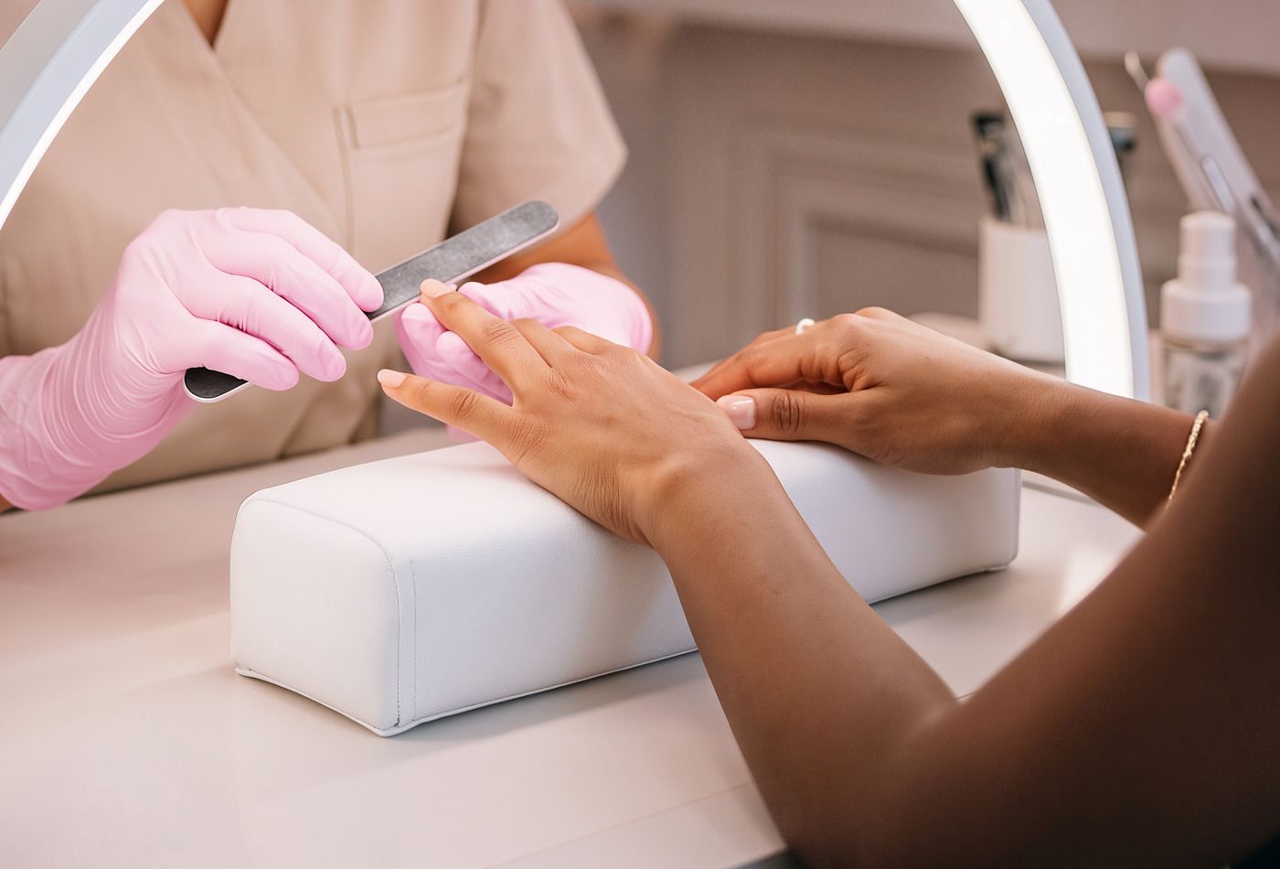 A client's hand rests on the white nail arm rest while a technician in pink gloves files a nail.